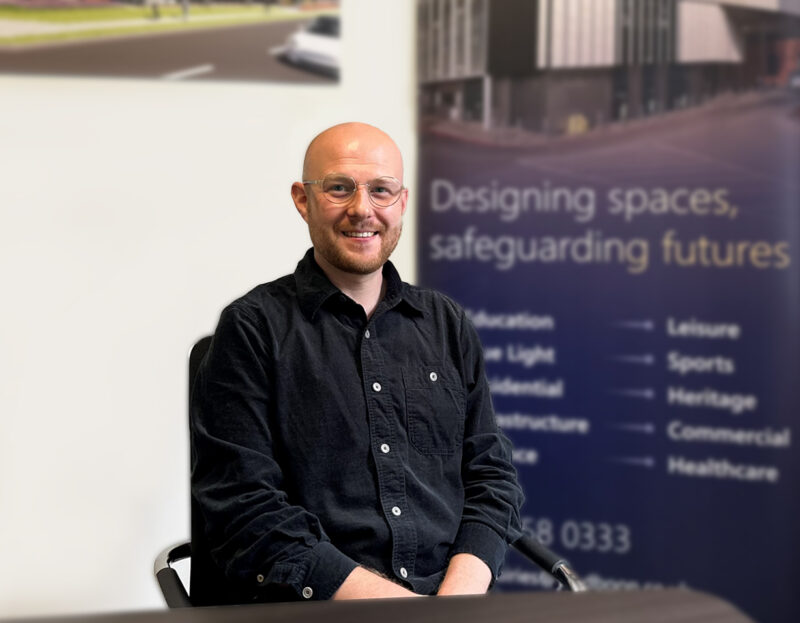 A man sitting in an office space, smiling at the camera. He is wearing a black shirt and glasses.