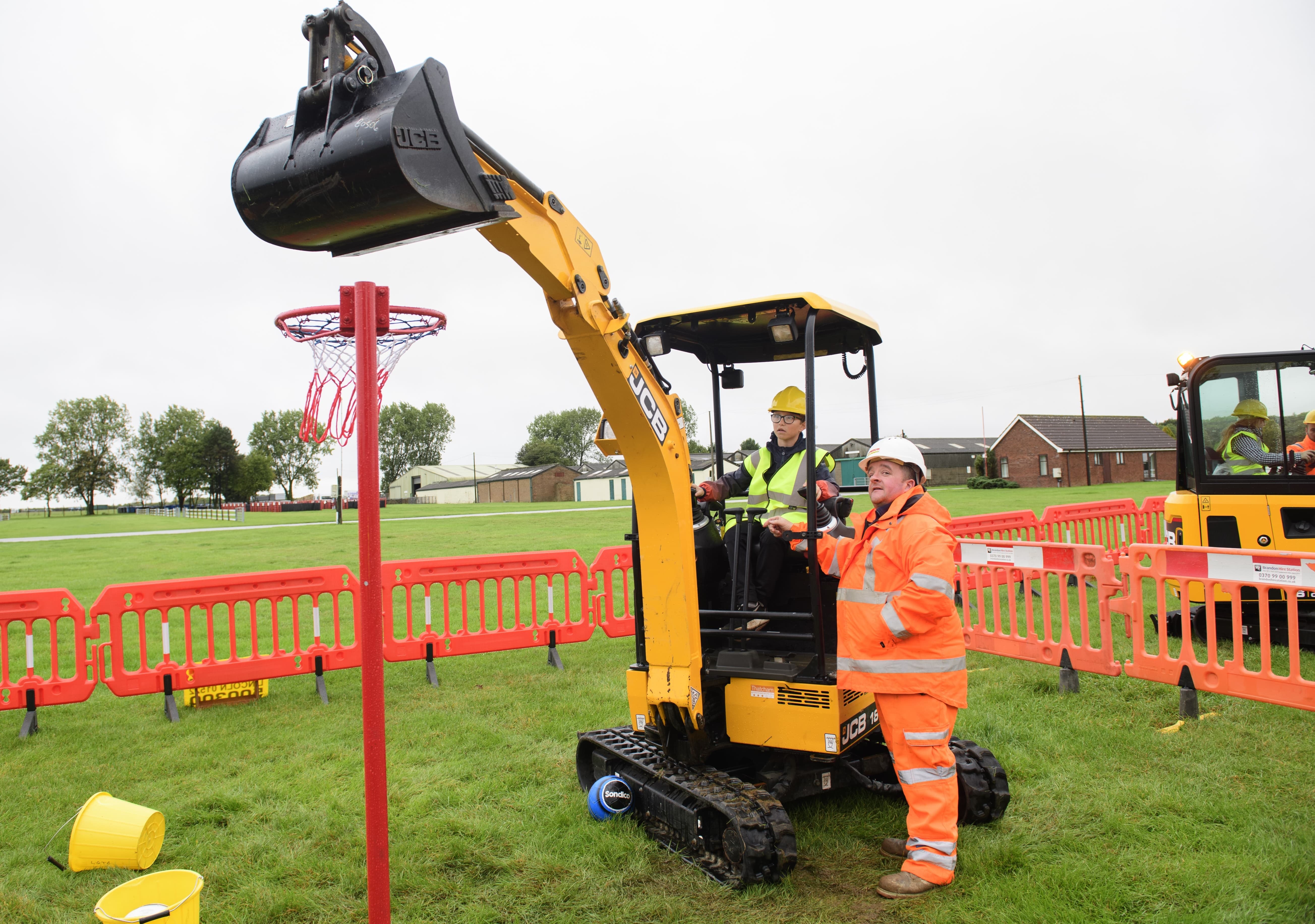 Young child operating a yellow JCB digger with man in orange high clothing supervising