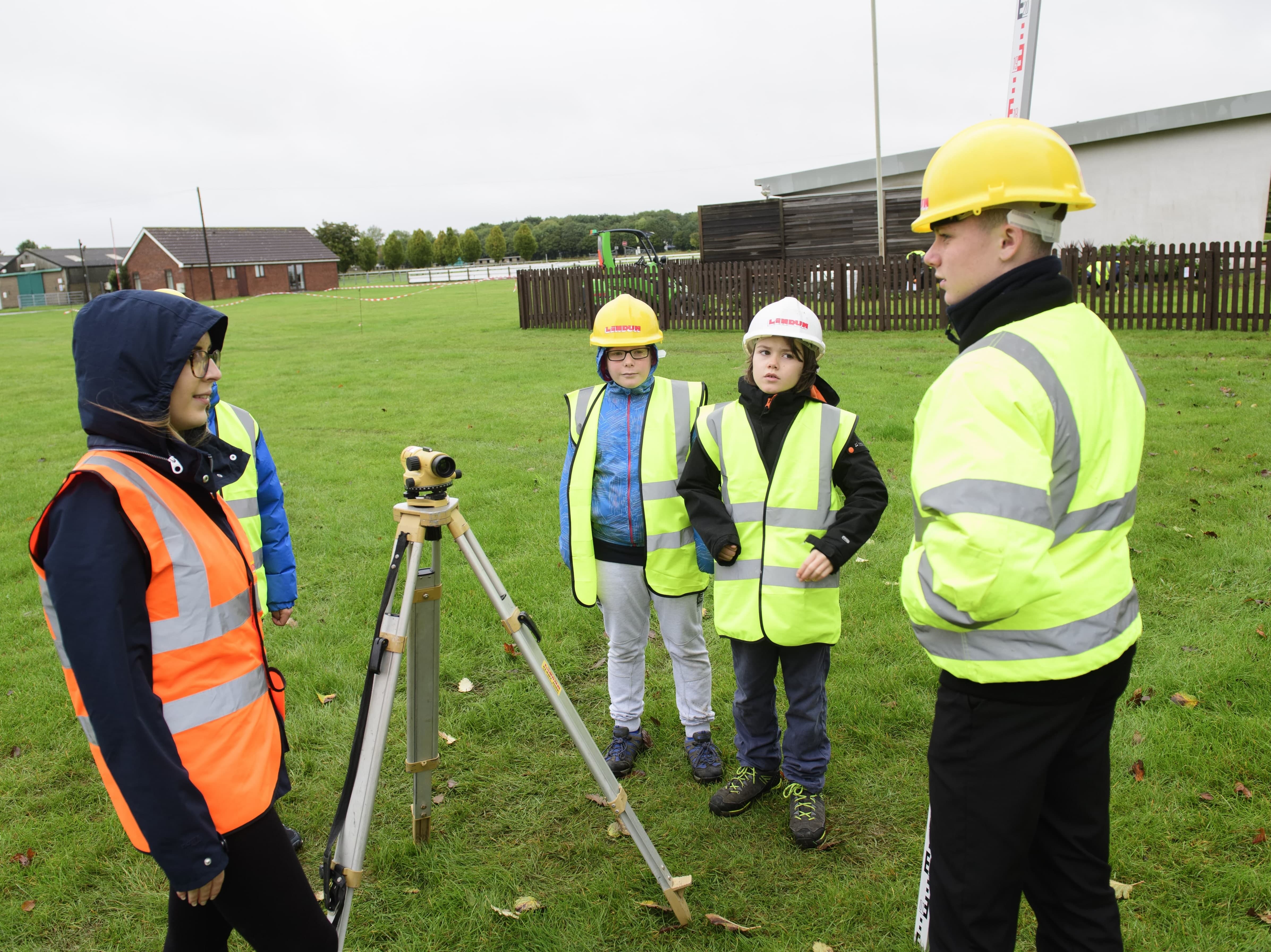 Young girl operating equipment with other children watching on, all wearing yellow high vis jackets