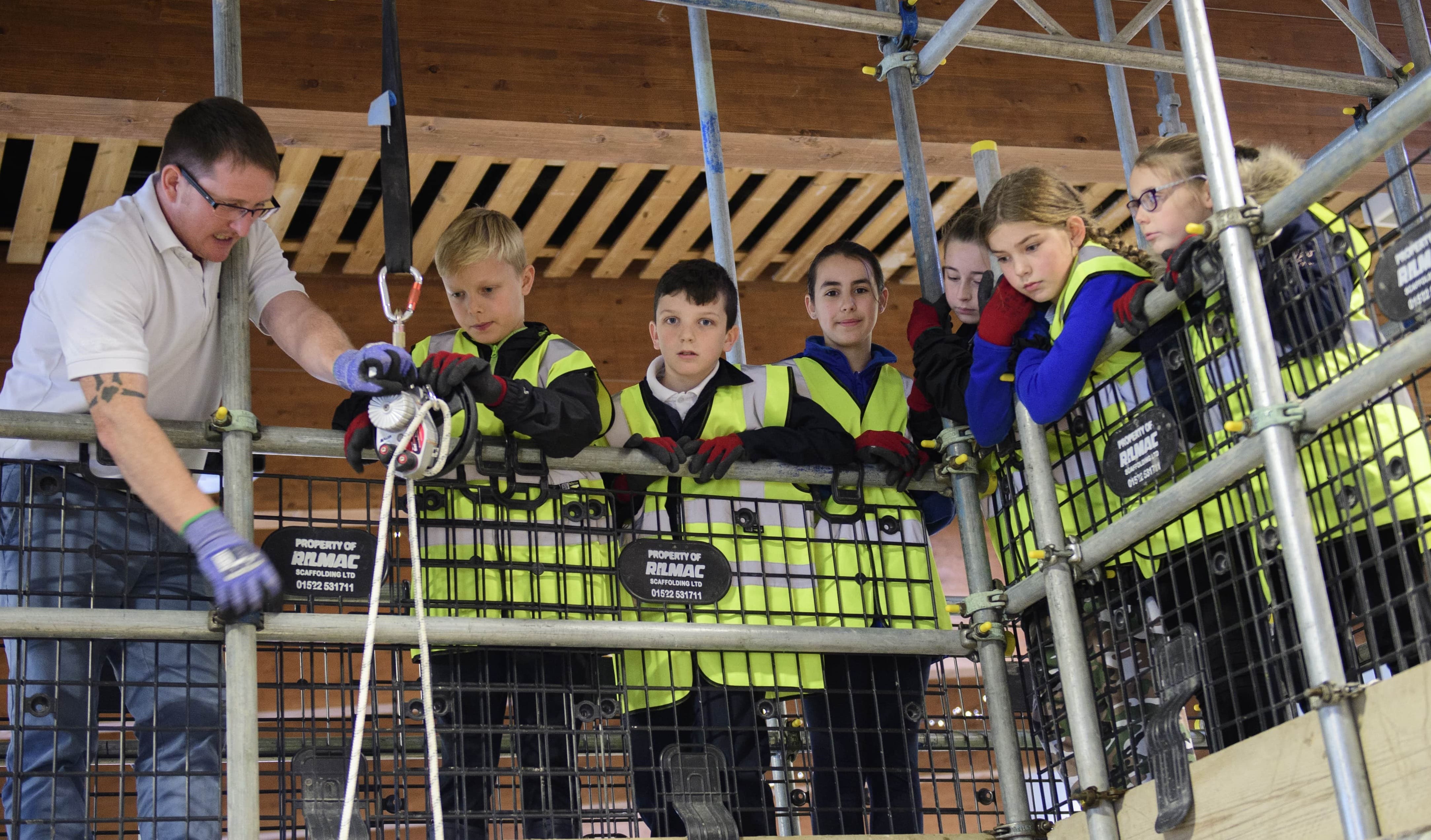 Young girl operating equipment with other children watching on, all wearing yellow high vis jackets