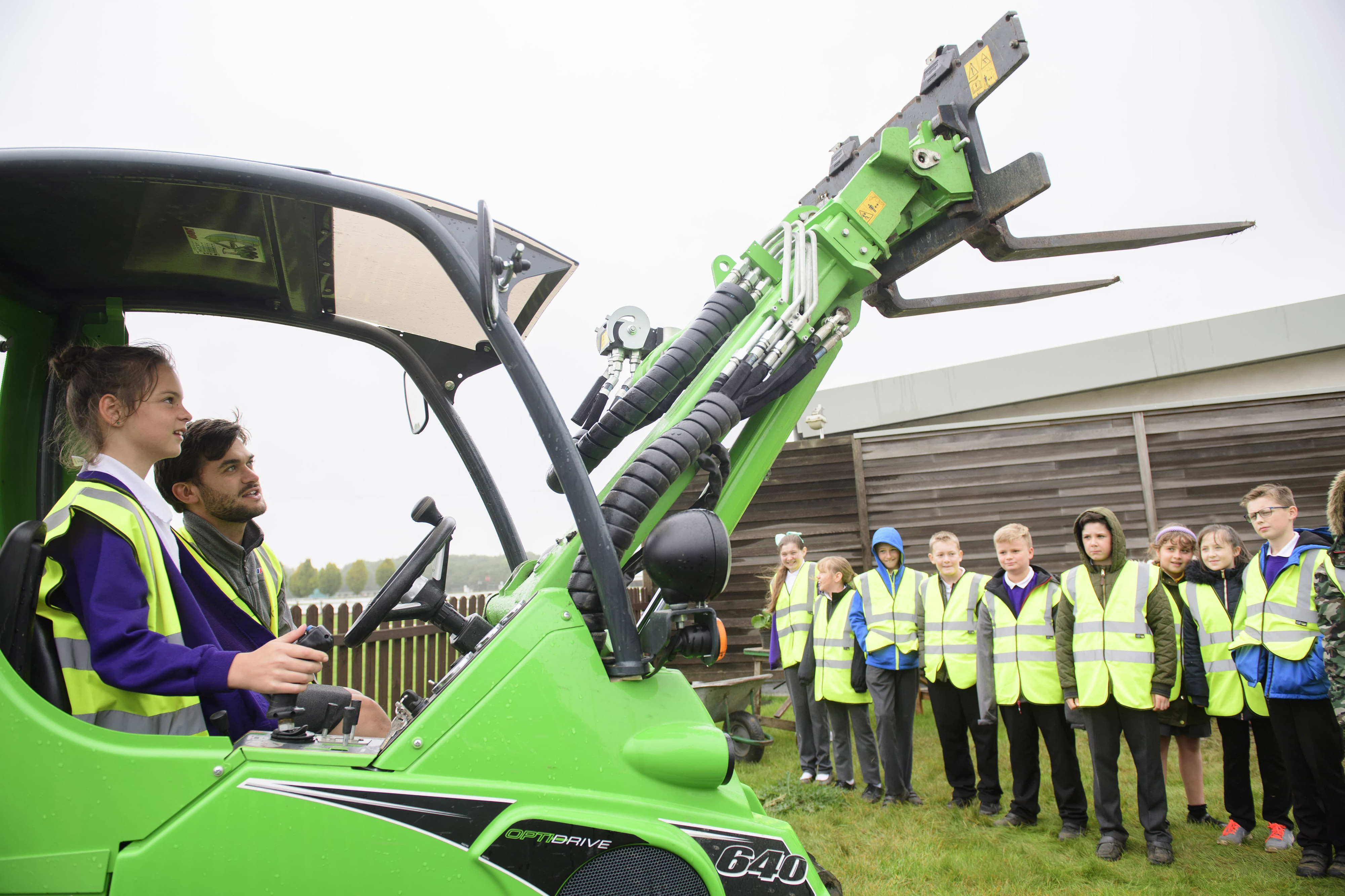 Young girl operating a green machine with other children watching on, all wearing yellow high vis jackets