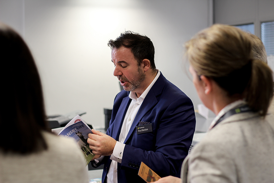 A man in blue suit jacket looking at brochure and speaking with others watching him