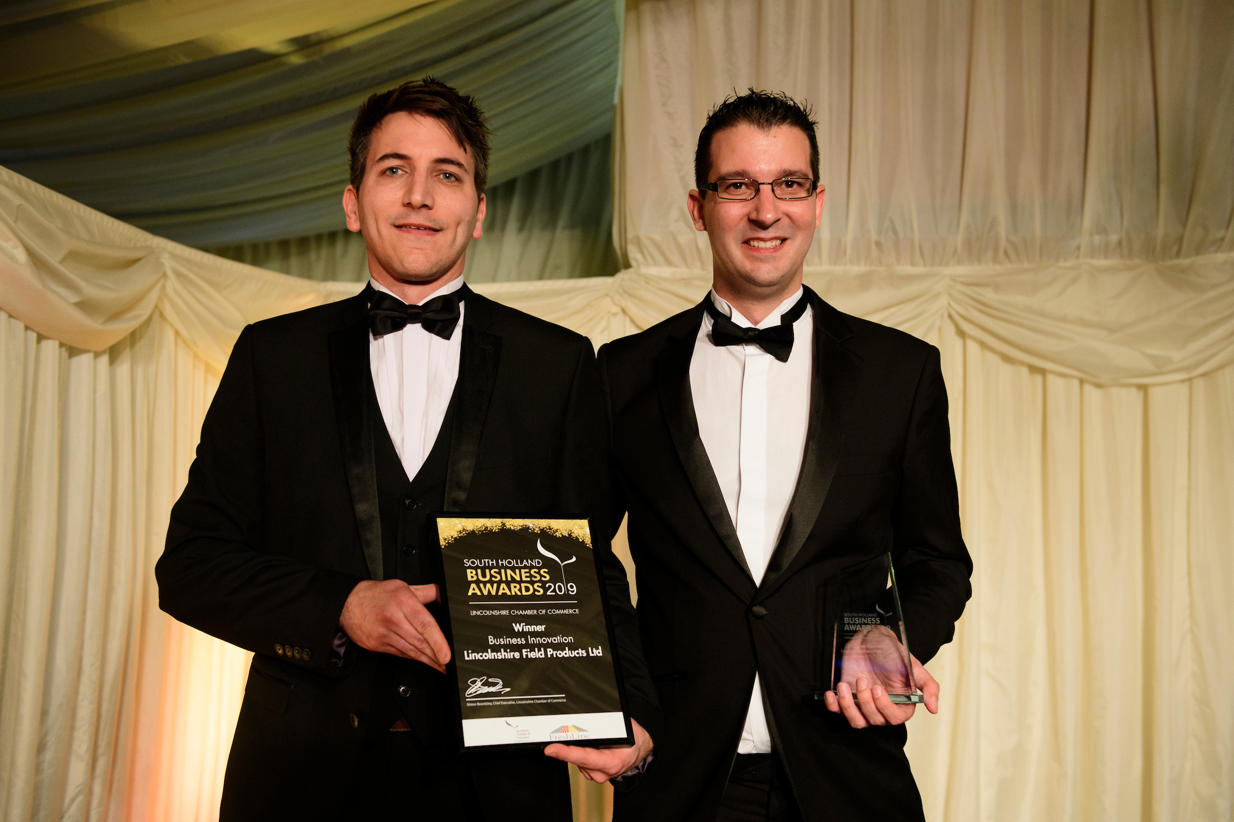 Two men in suits and bowties, one is holding a framed certificate and trophy, at the South Holland Business Awards 2019