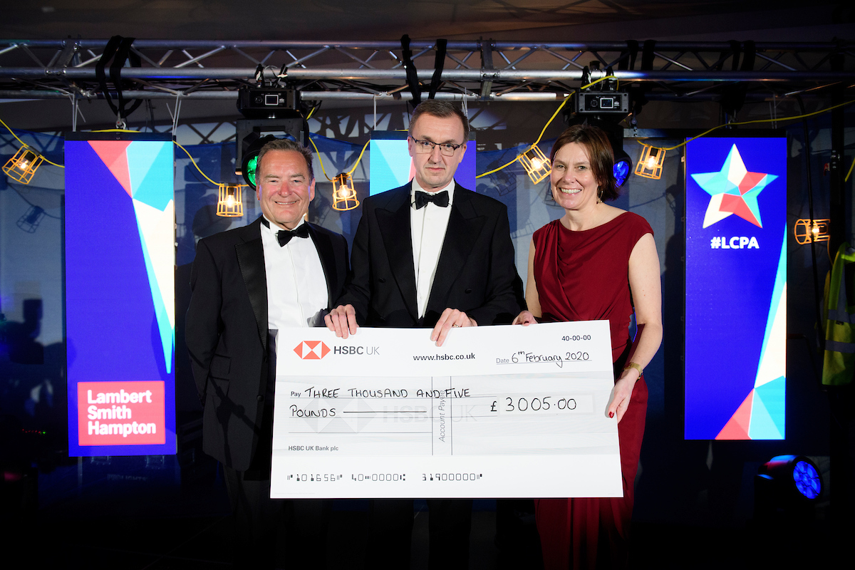 Three people in eveningwear, wearing bowties and suits with the lady in red, holding a large cheque