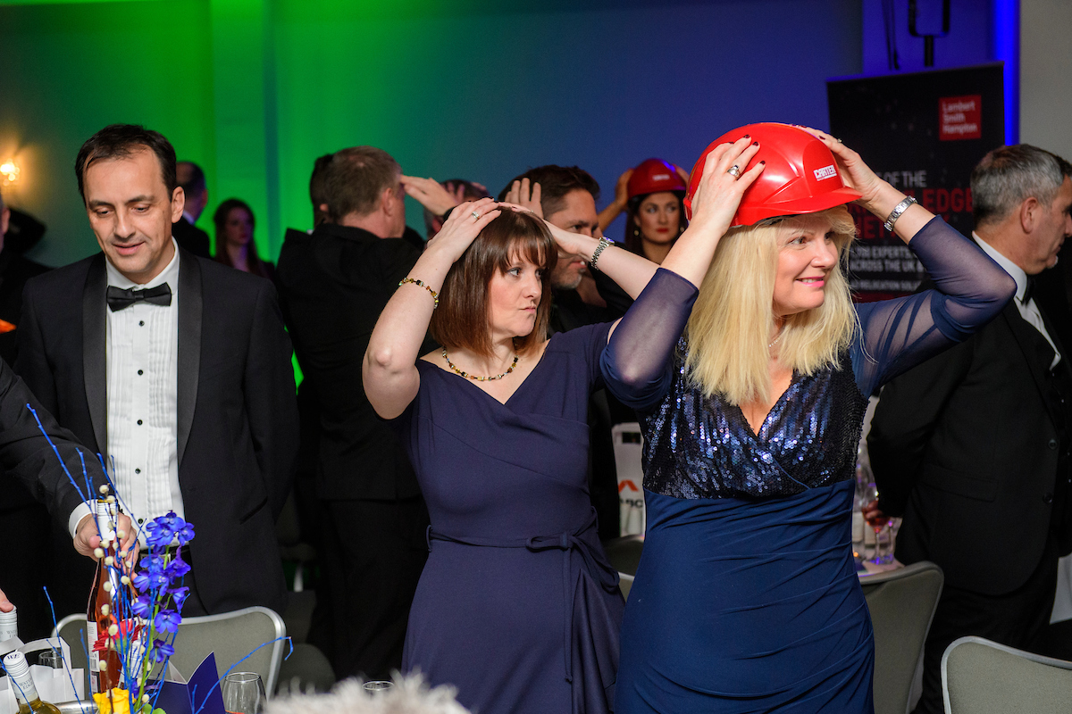 Several people wearing eveningwear and red hard hat standing up at an awards show with their hands on head