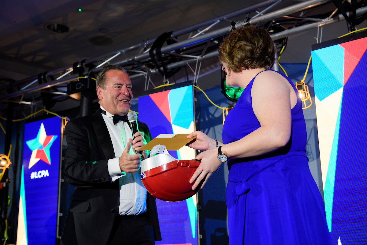 Woman with blue dress holding a red hard hat and gold envelope on stage at The Greater Lincolnshire Construction and Property Awards 2020 with host Jeff Stelling
