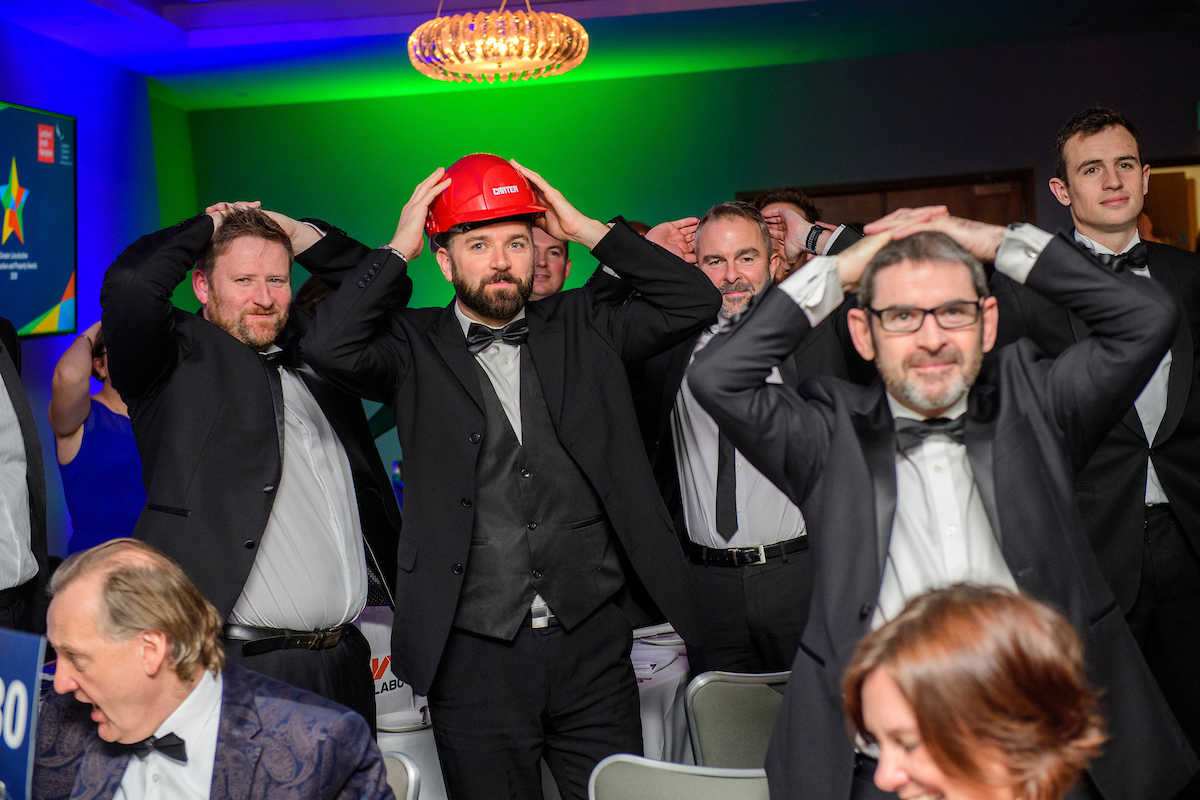 Several people wearing eveningwear and red hard hat standing up at an awards show with their hands on head