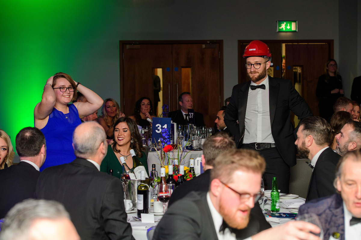 Young woman in blue dress and man wearing eveningwear and red hard hat standing up at an awards show