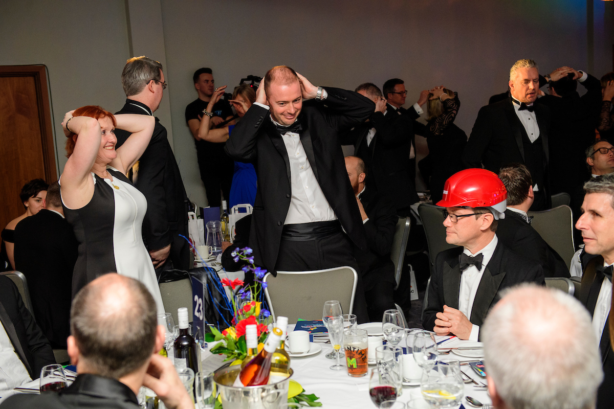 Several people wearing eveningwear and red hard hat standing up at an awards show with their hands on head
