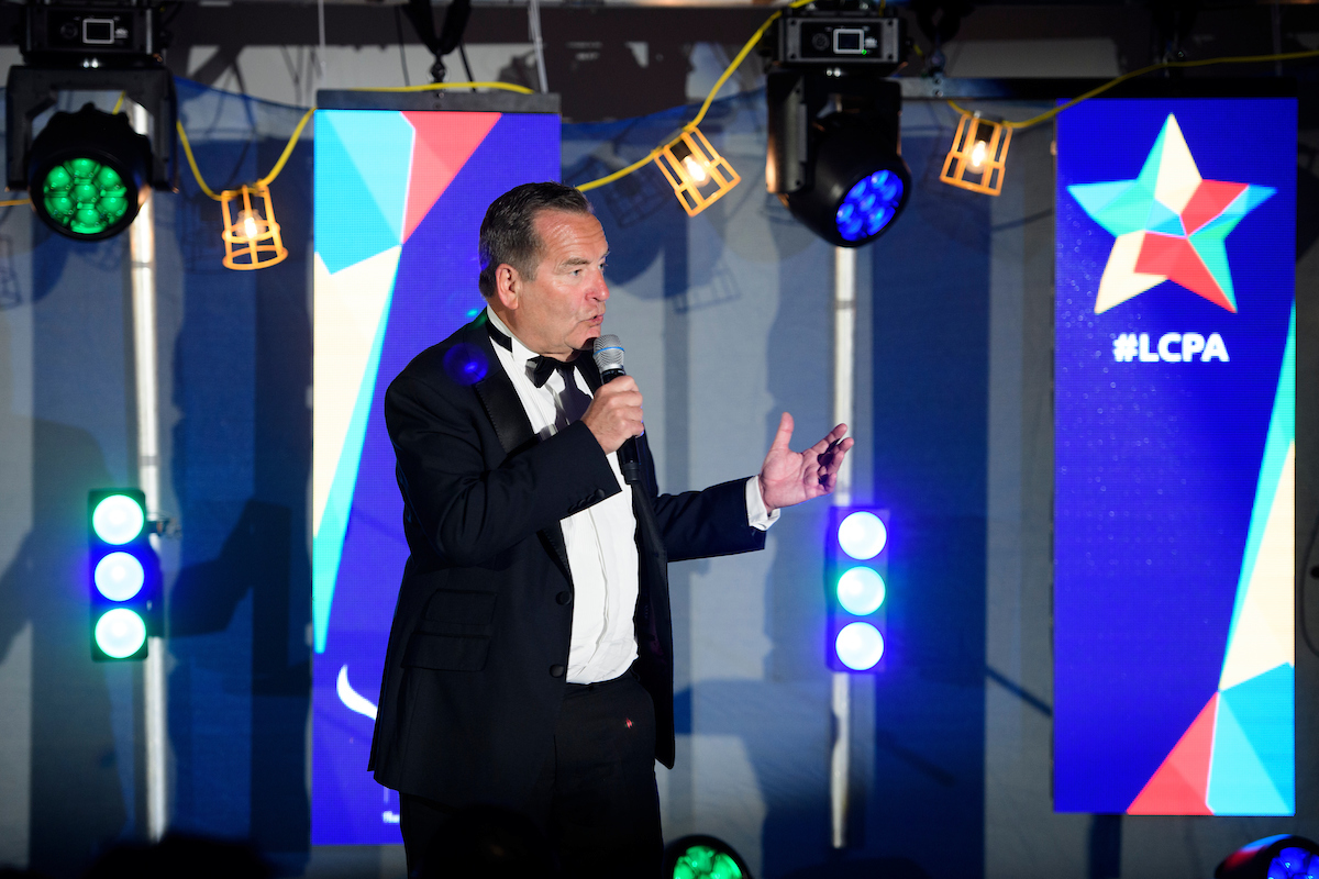 Picture of a man wearing a black suit and bowtie holding a microphone with digital screens showing blue background and #LCPA text