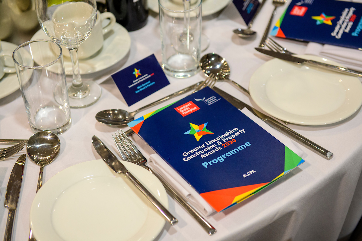 Picture of a set table with white table cloth, cutlery and plates, a blue booklet lies on top of one plate with text The Greater Lincolnshire Construction and Property Awards 2020