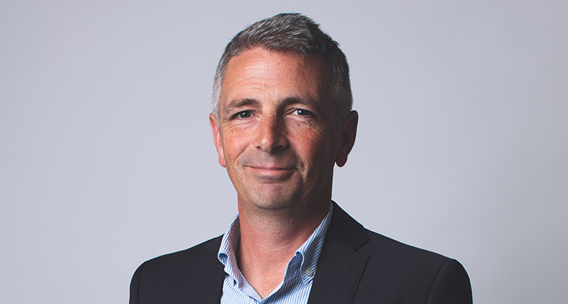 Picture of grey haired man smiling wearing a black jacket and blue and white striped shirt on grey background