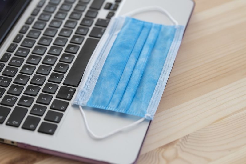 Picture of blue surgical face mask on top of light grey laptop keyboard on a brown table