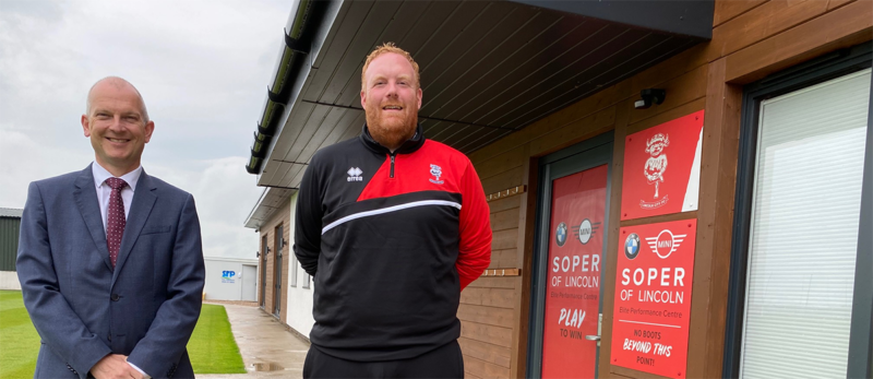 Two men posing and smiling in an outdoor setting in front of a door with red banners and Lincoln City FC Imp logo