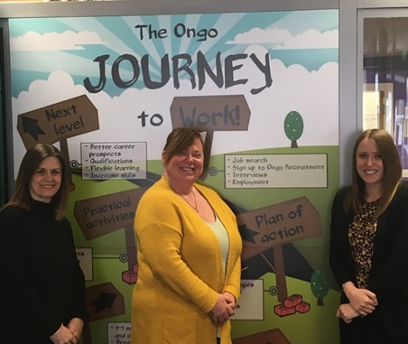 Three women, the middle one in a yellow dress, smiling in front of banner