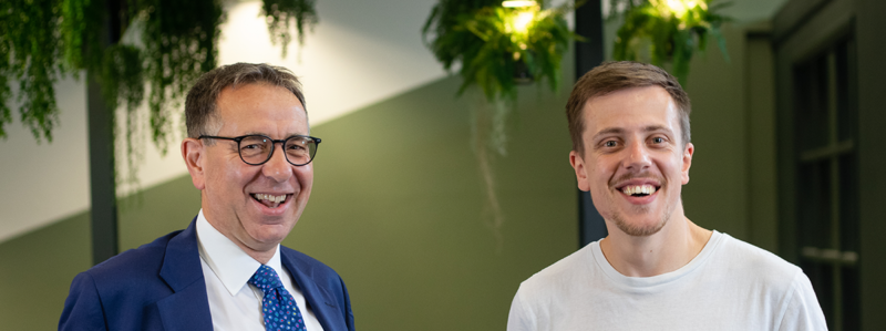 Two men smiling, the man on the left is James Pinchbeck of Streets Chartered Accountants, wearing glasses and a suit, the man on the right is Peter Watson of District, wearing a white T-shirt