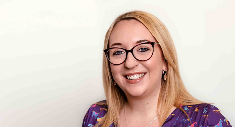 Young woman with long blonde hair, glasses and purple top smiling