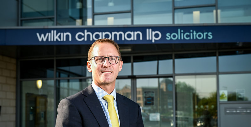 Robin Simmonds, CEO of Wilkin Chapman. Man wearing suit with yellow tie, wearing glasses, in front of office exterior