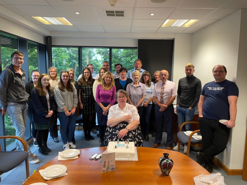 A group of men and women standing behind a woman sitting on a chair in front of a table with cake, in an office setting