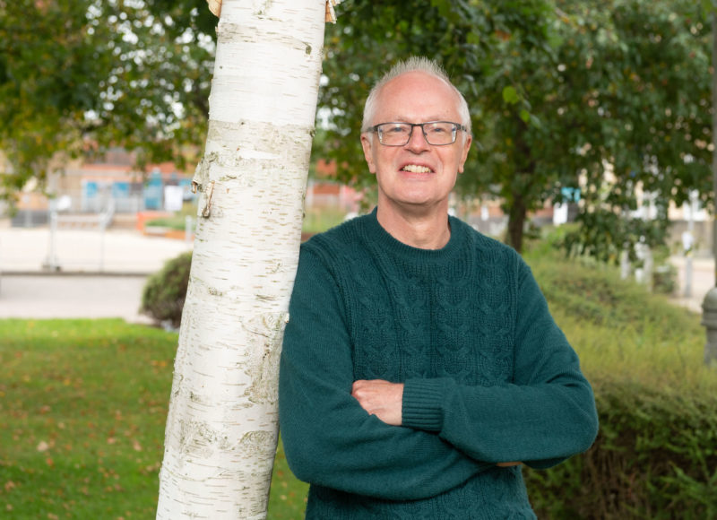 Barrie Billinghay, a man with short grey hair, wearing glasses and a dark green jumper in outdoor setting leaning against a tree