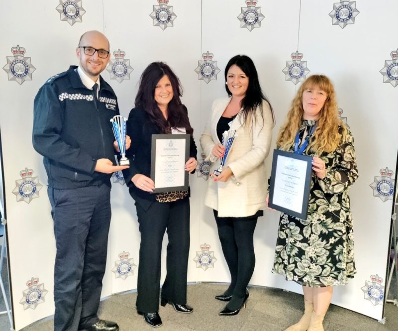 Police officer with three women, holding trophies and certificates on background with Humberside Police logos behind them