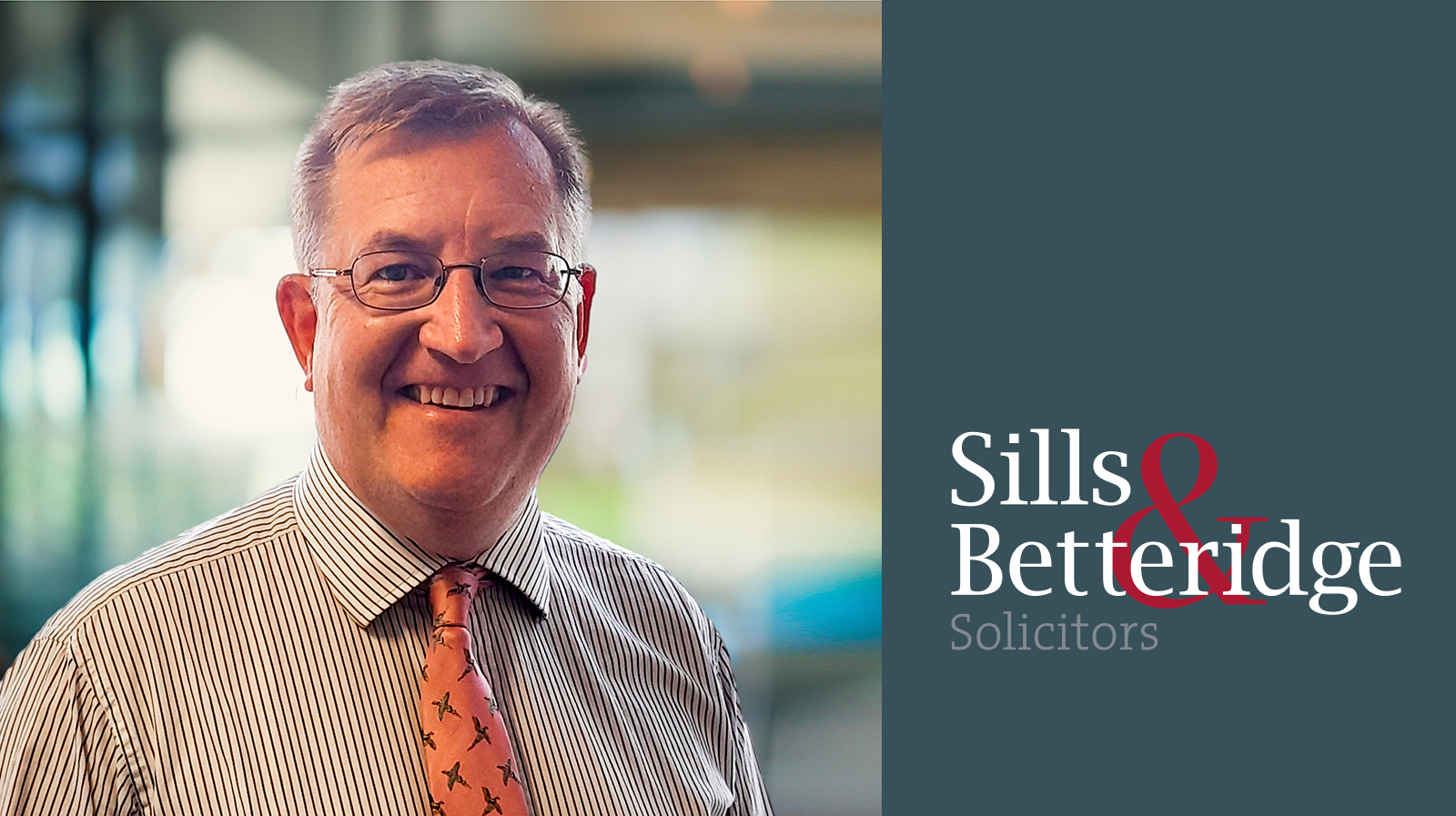 David Wood, partner at Sills & Betteridge, a man wearing glasses, striped shirt and bright tie, next to grey background with Sills and Betteridge logo in white and red