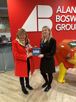 Two women holding a plaque in front of red banner with Alan Boswell Group logo