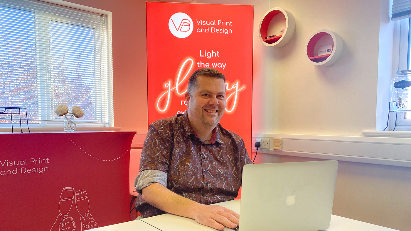 Man wearing patterned dark red shirt in office with red banner showing Visual Print branding behind him. He is sitting at a white desk using a silver laptop