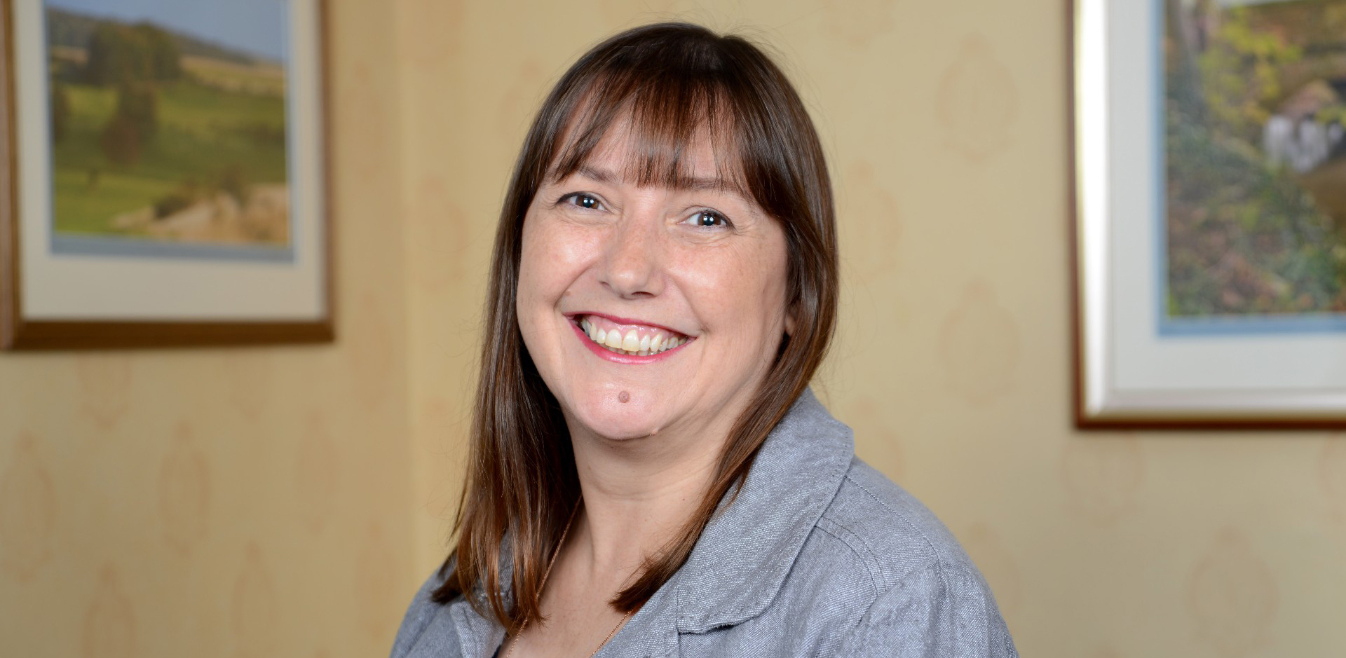 Woman with shoulder length dark hair, grey jacket, smiling at the camera with beige background