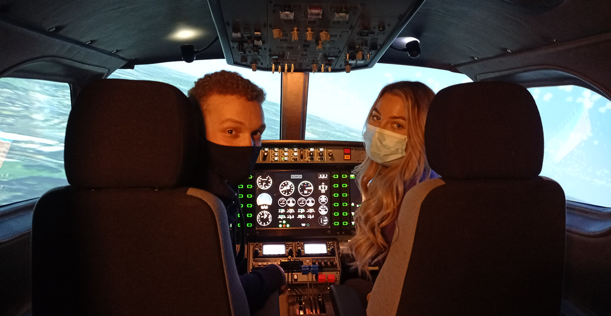 Young man and woman wearing face masks sitting in pilot seats within a plane's cockpit