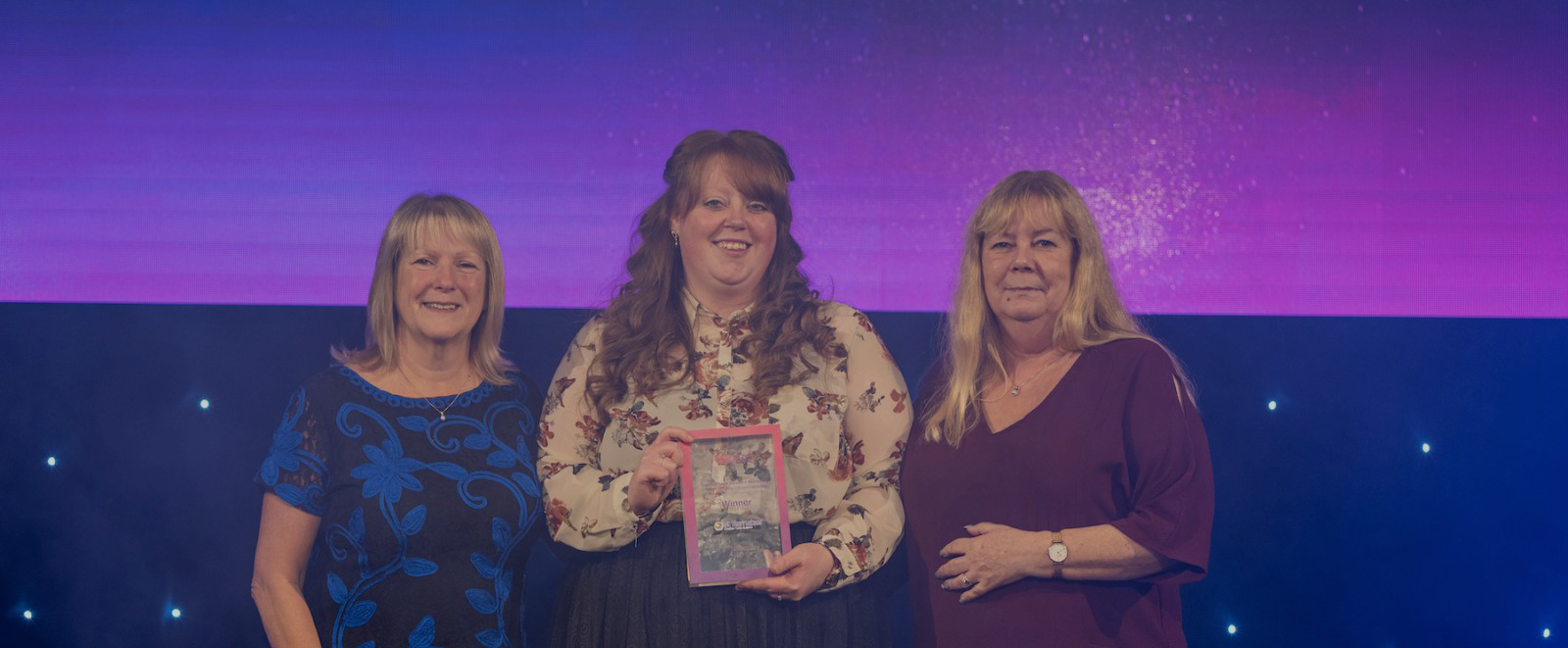 Three women in evening dresses holding trophy at award show with purple lighting