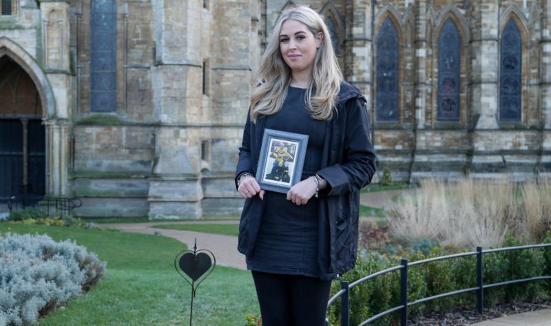 Young woman with blonde hair holding a framed picture of her father standing next to an iron heart by the Lincoln Cathedral