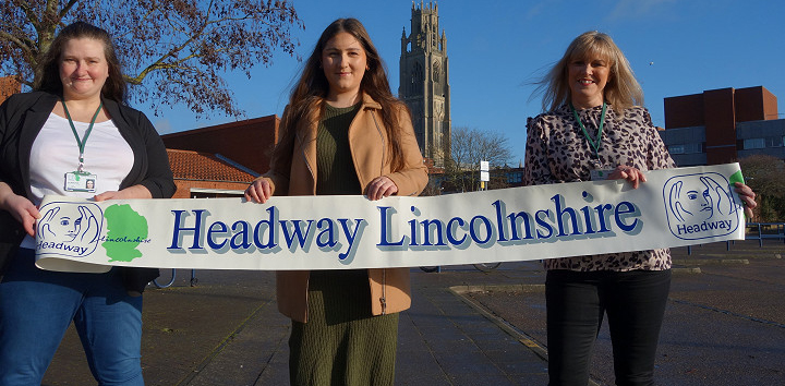 Three woman outdoors holding long white banner with Headway Lincolnshire logo