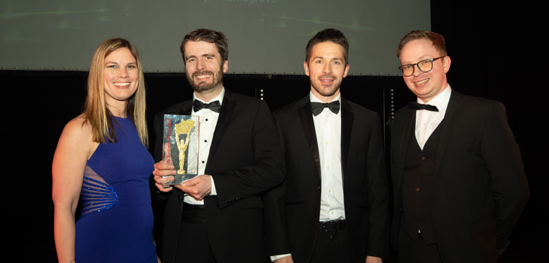 Blonde woman in blue dress with three men dressed in black suits at awards ceremony. DBS Team receiving Midlands Award, Service Provider of the Year (l-r) Julie Priestley, James Hopkins, Matt Rayner & Dan Day