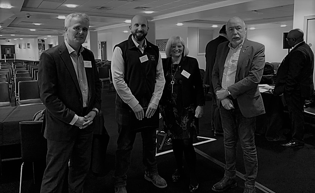 Black and white picture of three men and a woman dressed in business attire at a conference