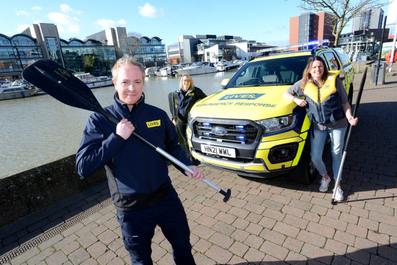 White male in the foreground holding rowing paddle with two woman in the background with yellow high visibility car with LIVES logo. They are at the Brayford marina in Lincoln, with water and buildings visible behind them.
