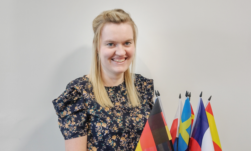 Smiling young blonde woman against grey background with flags from different countries.