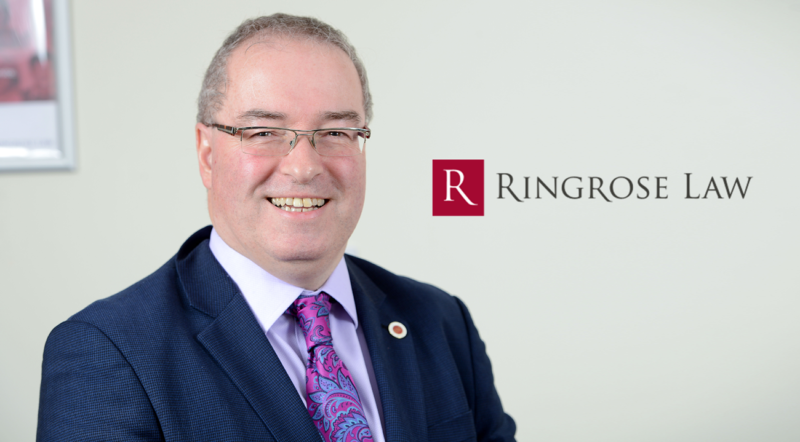 White male wearing glasses and dark blue suit jacket, purple tie, on grey background smiling, with Ringrose Law logo in red and grey