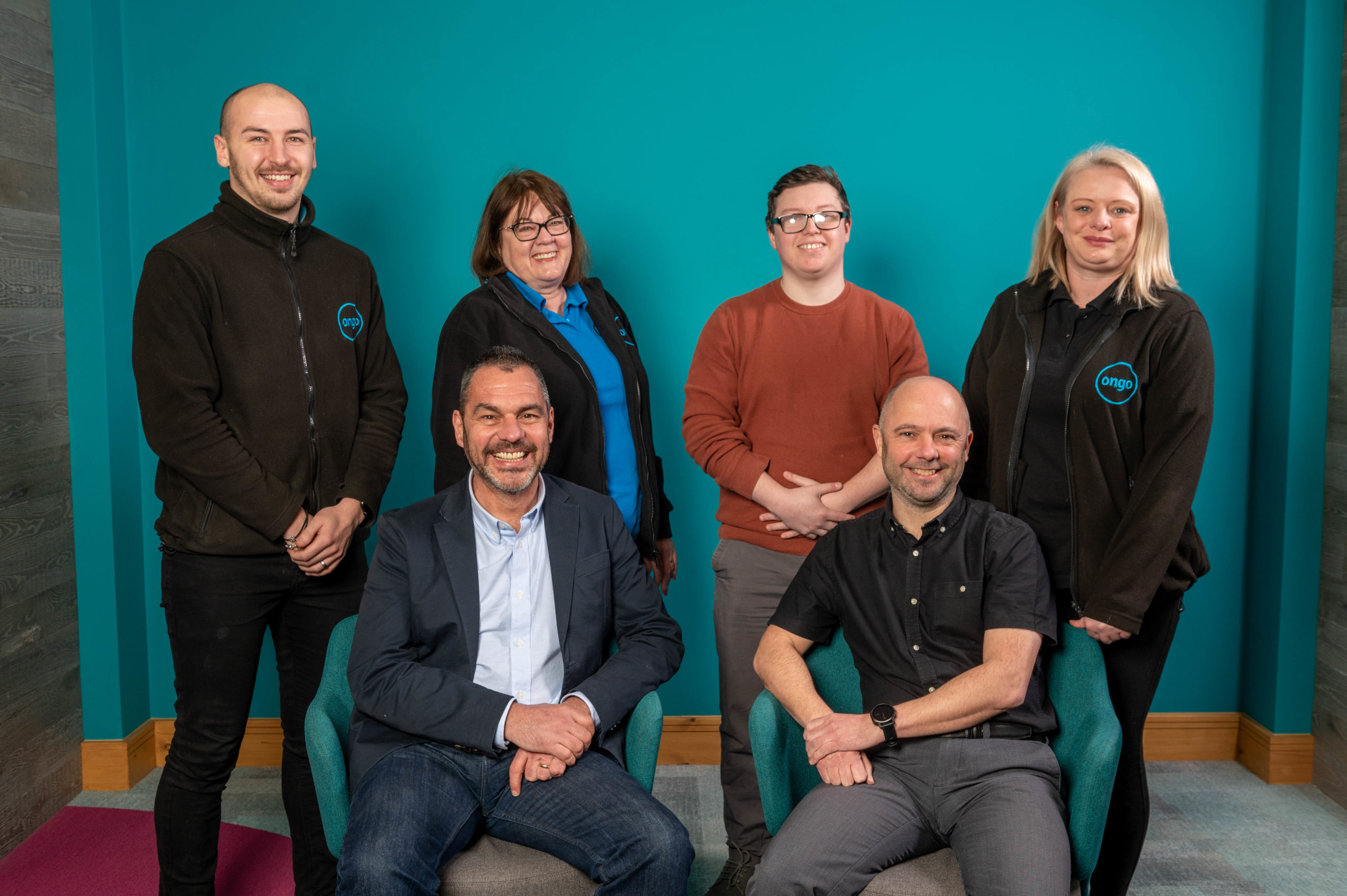 Group of men and women sitting and standing in front of teal background, smiling at camera