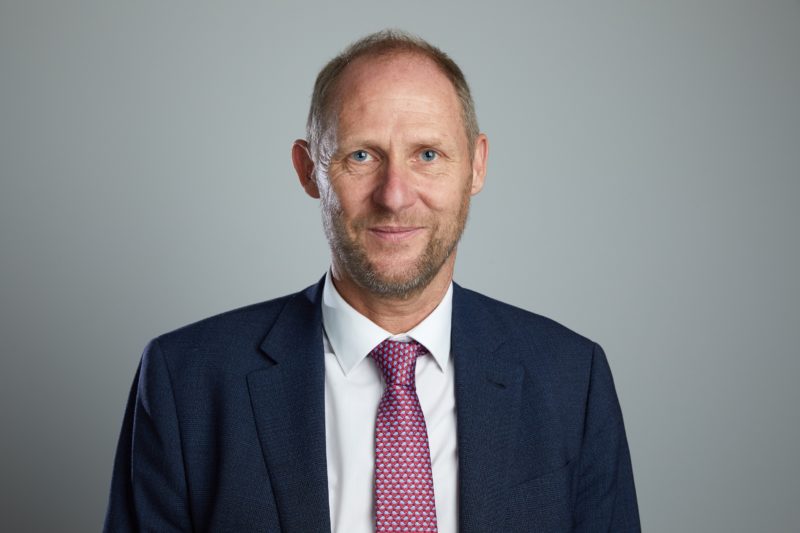 White male with short hair and beard, wearing dark blue suit jacket, white shirt and red tie