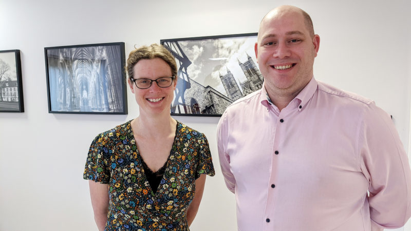 Woman with hair in ponytail wearing glasses and floral dress, next to man in light pink shirt, both smiling at the camera with pictures of Lincoln Cathedral in the background on a wall.