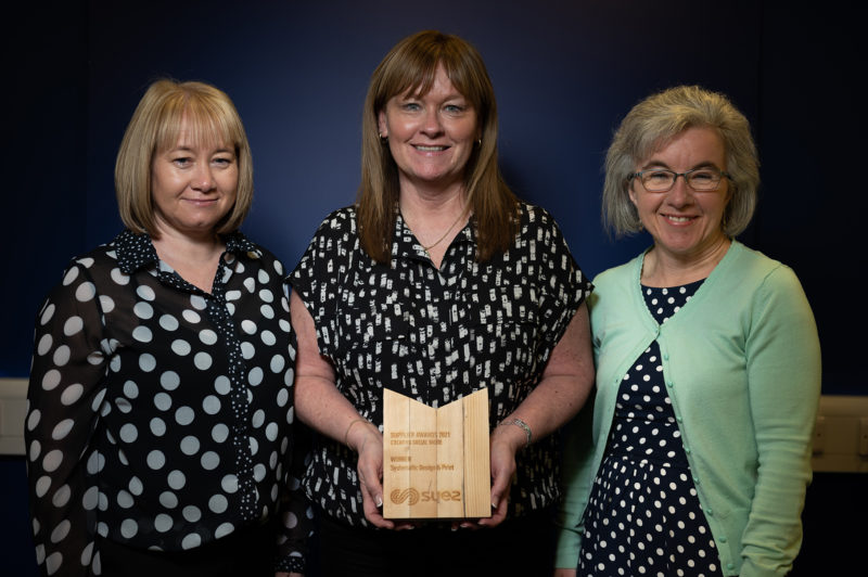 Three women with the middle holding a wooden trophy with text "Supplier awards 2021, creative social value, winner Systematic Print Design, SUEZ". The women are against a dark blue backdrop