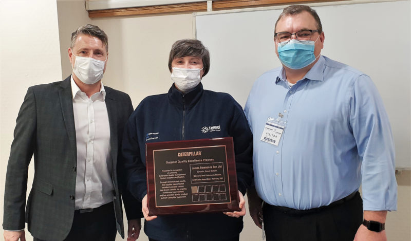 Two men and a woman holding a wooden award. From left to right: Man in grey suit with white shirt, woman with branded dark blue fleece jacket holding trophy, man with blue shirt and black trousers. All are wearing facemasks.