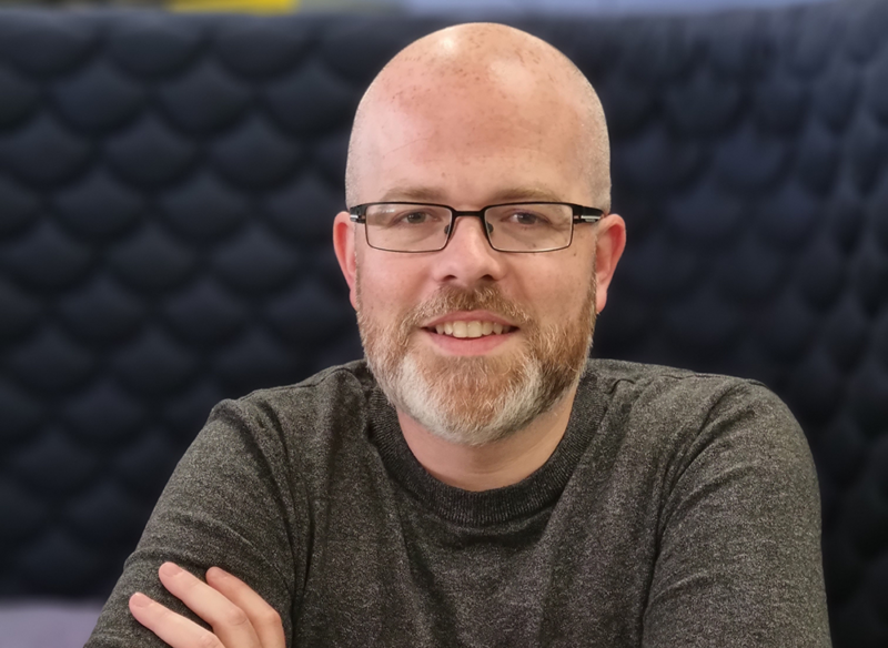 Bald white male with salt and pepper beard and wearing black glasses and grey top, smiling at camera