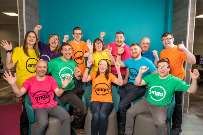 Group of people standing and sitting, wearing brightly coloured T-shirts with black Ongo logo, against bright blue background