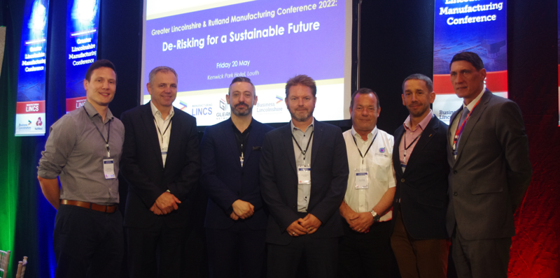 Group of seven white males posing at the Business Lincolnshire Manufacturing Conference 2022, with presentation on a screen behind them, branded banners, and red, green and blue lighting.