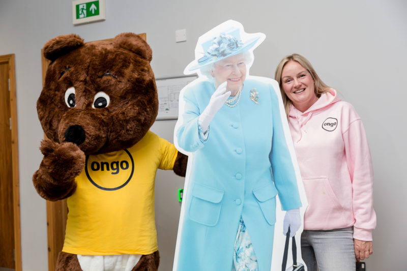 Cardboard cut-out of Queen Elizabeth II dressed in blue, with a person dressed in bear mascot costume wearing yellow T-shirt with Ongo logo, and woman wearing pink, behind the cutout.