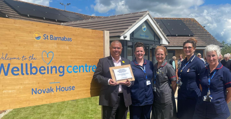 Professor Sir Jonathan Van-Tam holding a plaque commemorating the opening of Novak House. Next to him are four women in nurse's outfits, and a large wooden sign next to them reads "St Barnabas. Welcome to the Wellbeing Centre Novak House" with exterior of building in background