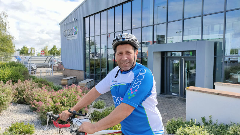 Man wearing bicycle helmet and blue and white lycra top on bicycle in front of APSS building with glass windows.