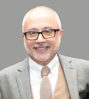 A man, wearing a grey suit, with glasses and a beard, smiling at the camera.